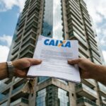 Leilão de imóveis Caixa: mãos segurando edital com o logo da Caixa diante de prédio residencial
